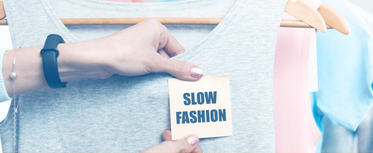 Mão feminina colando um post-it que tem escrito "slow fashion" em uma blusa estilo regata de cor cinza.