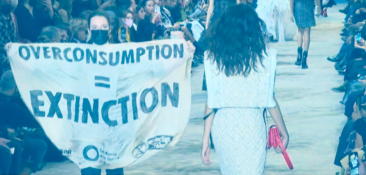 Foto de um protesto em um desfile de moda, com uma placa escrito "Consumo desenfreado igual a Extinção"